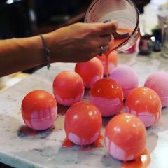 Noelle, our creator, pouring orange cocoa butter glaze onto pink bath bombs on a marble counter